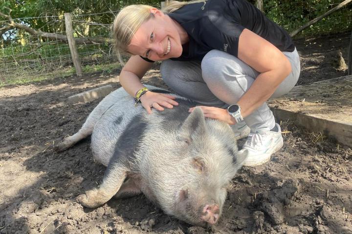 Woman crouching and smiling next to a sleeping pig in a muddy outdoor area with trees.
