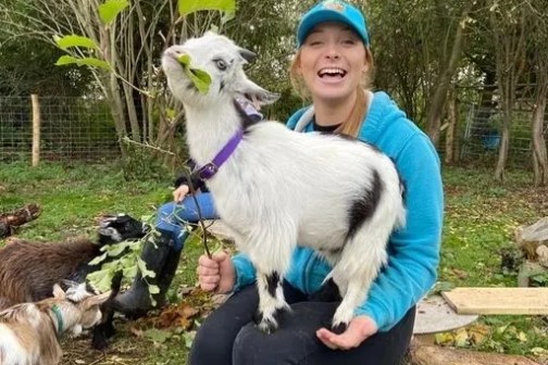 Person in blue outfit sitting with a small goat on lap in a wooded area.